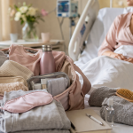 Gepackte Kliniktasche mit Kleidung und Pflegeprodukten im Krankenhauszimmer vor einer Patientin im Bett