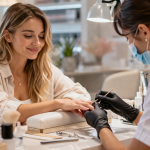 Frau bekommt Maniküre im Nagelstudio während die Nageldesignerin eine Schutzmaske trägt – Hinweis auf chemische Belastung bei Nagelpflege
