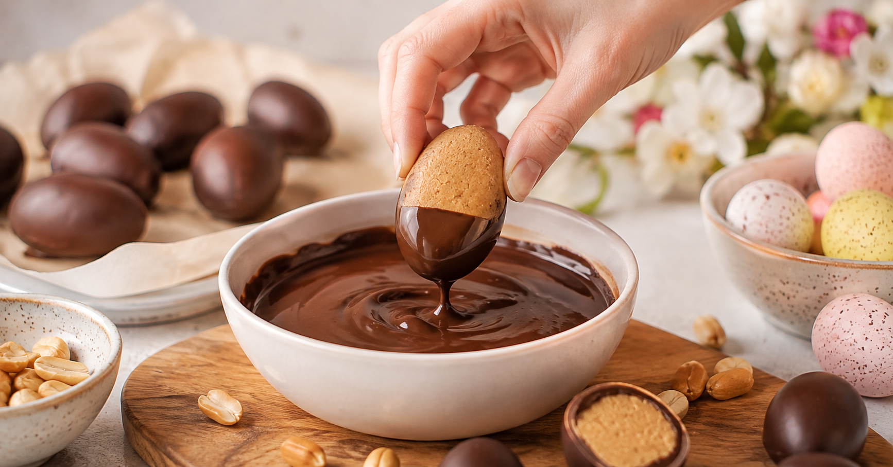 Eine Hand taucht ein selbstgemachtes Peanut Butter Easter Egg in geschmolzene Zartbitterschokolade. Im Hintergrund liegen fertige Schoko-Erdnussbutter-Eier und Osterdekoration.